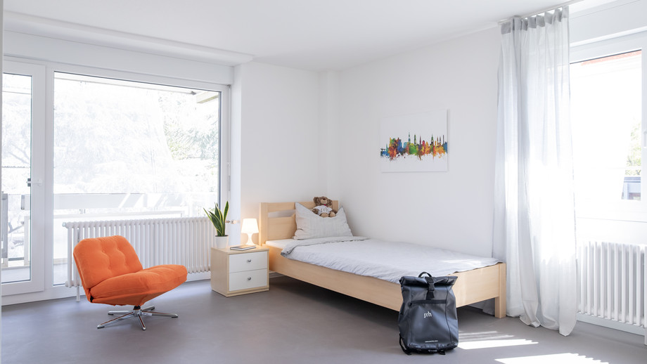 Ein helles Schlafzimmer mit Bett, Nachttisch, orangefarbenem Sessel, Pflanze und einem schwarzen Rucksack am Boden. Große Fenster mit Vorhängen und ein abstraktes Bild an der Wand.