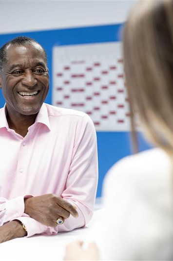 Ein älterer Mann lächelt freundlich in einem Büro, während eine Person ihm gegenüber sitzt und eine Frau im Hintergrund arbeitet.