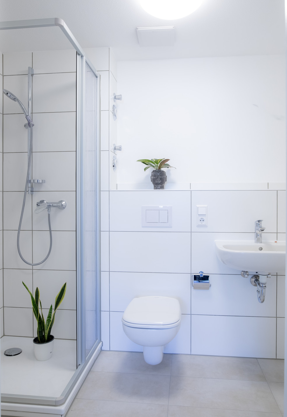 Badezimmer mit weißer Fliesenwand, Dusche mit Vorhang, WC und Waschbecken, zwei Pflanzen als Deko.