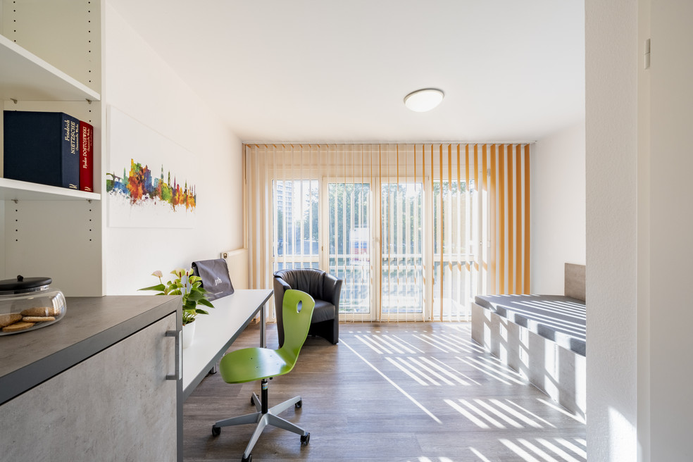 Moderner Arbeitsbereich mit Schreibtisch, grünem Drehstuhl und Blick auf Balkon mit Glasdoor. Bücherregal und Pflanze links im Raum.