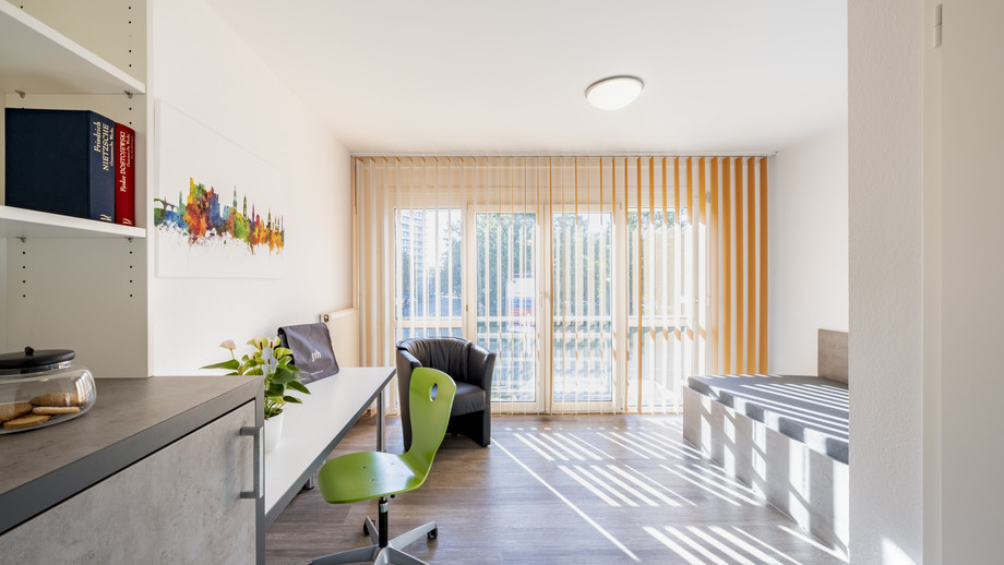 Moderner Arbeitsbereich mit Schreibtisch, grünem Drehstuhl und Blick auf Balkon mit Glasdoor. Bücherregal und Pflanze links im Raum.