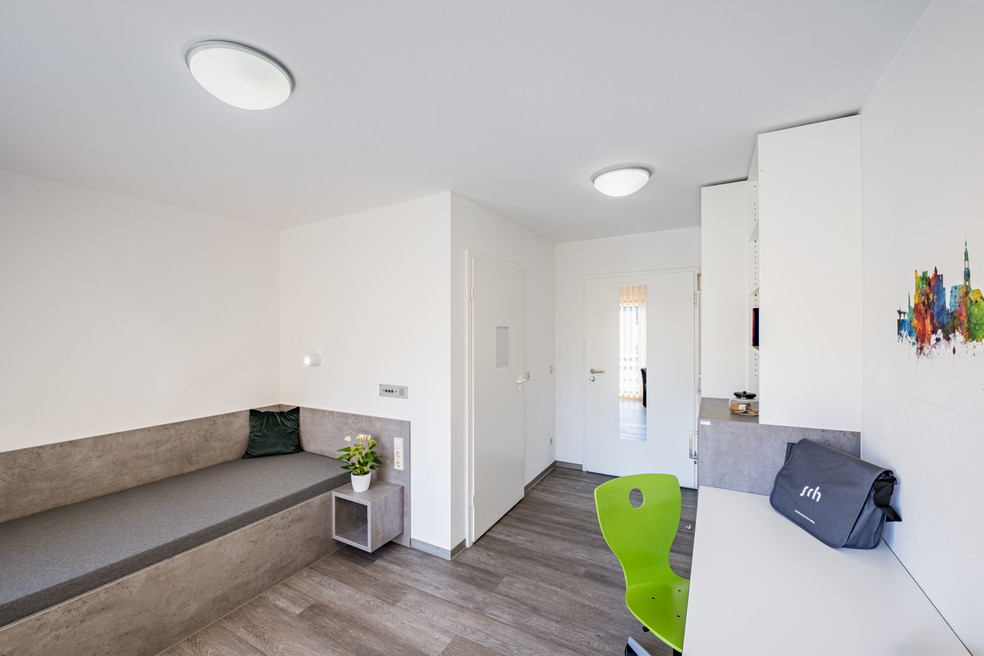 Moderner Wohnraum mit grauer Sitzbank, grüner Hocker, Pflanze und Tür zum Flur.