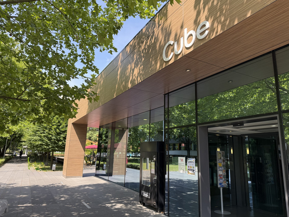 Moderne Gebäude-Fassade mit dem Schriftzug Cube, gläserne Eingangstür und grüne Bäume im Vordergrund.