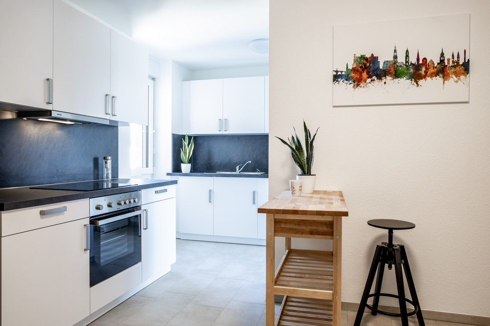 Moderne Küche mit schwarzer Arbeitsplatte, weißer Einbauküche und Herd. Daneben kleiner Tisch mit Pflanze und Hocker sowie ein Stadtgemälde an der Wand.