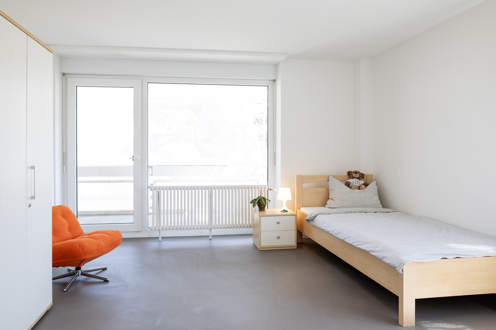 Ein helles Zimmer mit Bett, Nachttisch, orangefarbenem Sessel und großer Fensterfront zur Terrasse. Ein Radiator steht neben dem Bett.