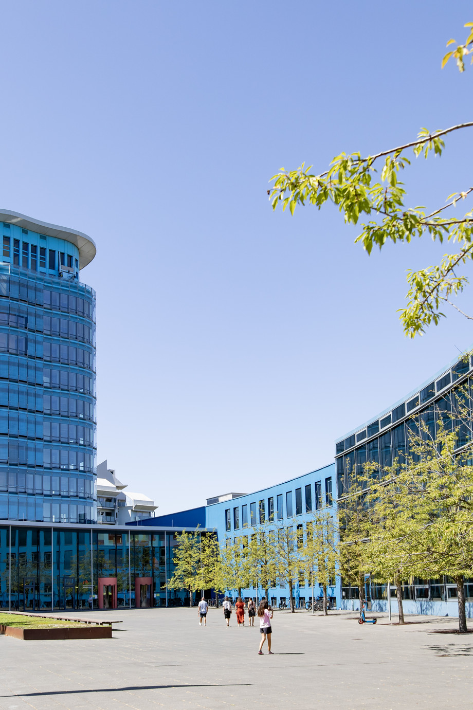 Moderne Glasgebäude mit runder Turmstruktur auf einem Platz, Flagge weht im Wind, Bäume und Menschen im Vordergrund.