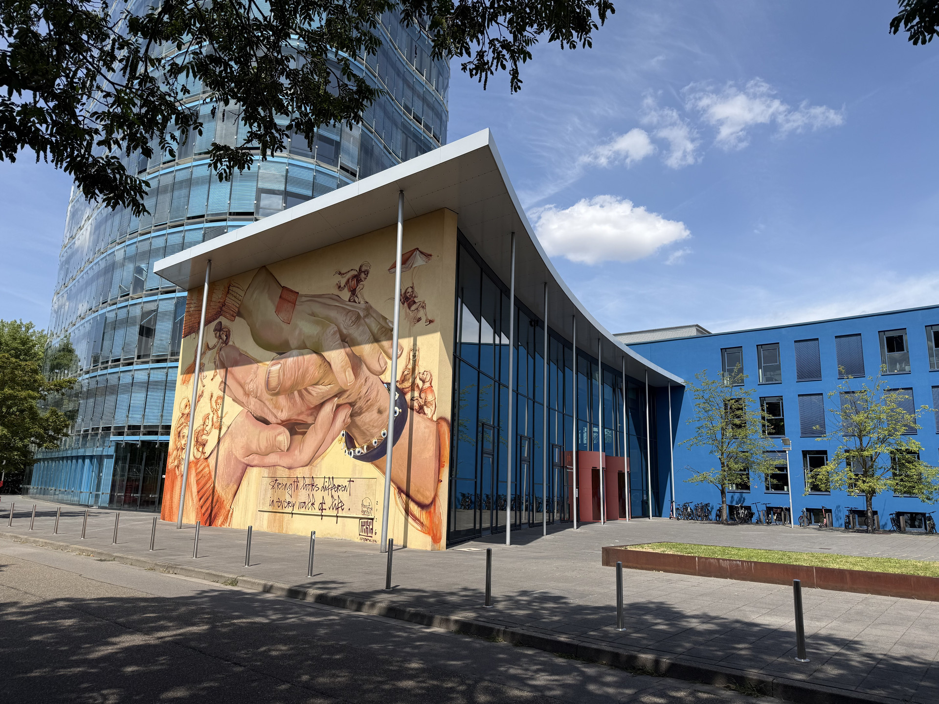 Moderne Gebäudeansicht mit großer Wandmalerei und Glasfront, daneben blaue Fassade mit Fahrradständern.