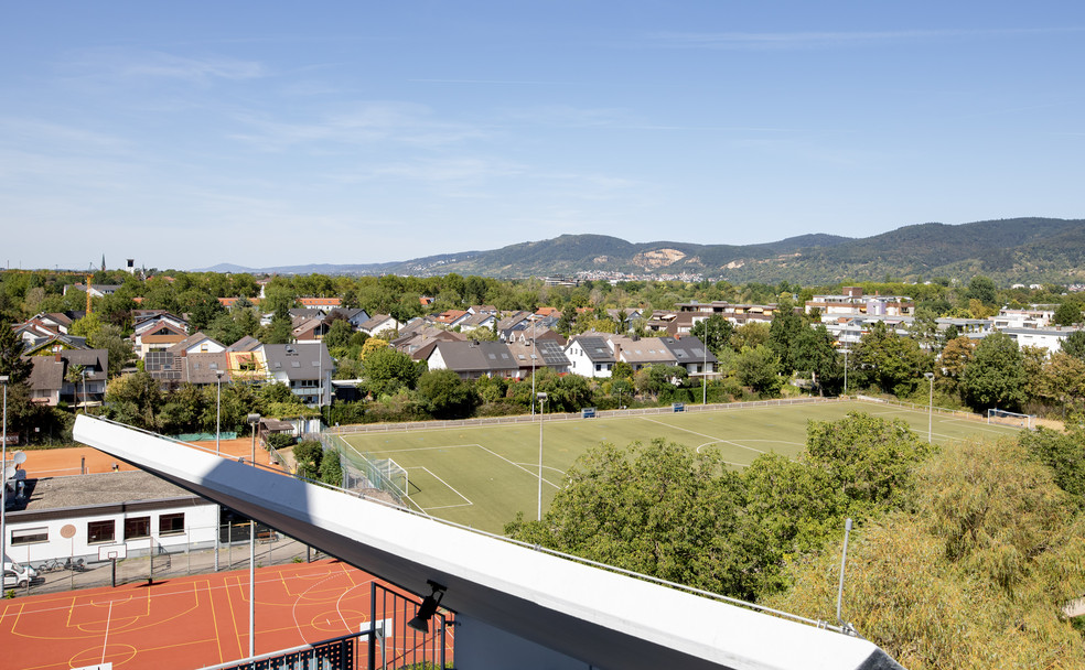 Panoramablick auf ein Wohngebiet mit Fußballfeld und Bergen im Hintergrund, klarer Himmel.