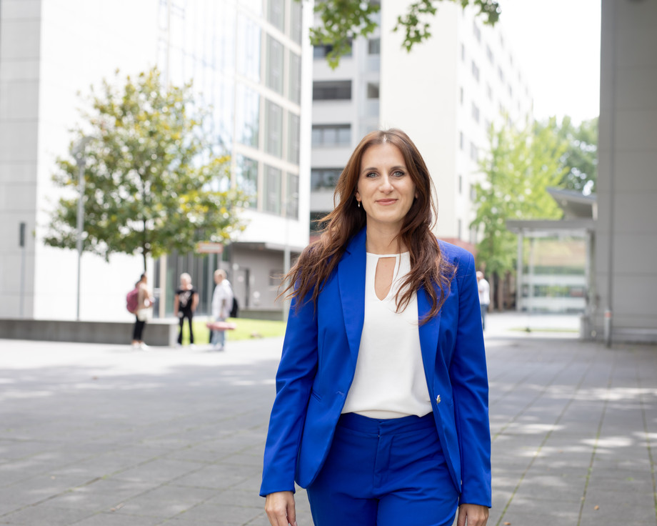 Eine Frau in blauem Anzug mit weißem Top steht lächelnd vor einem Gebäude mit Bäumen im Hintergrund.