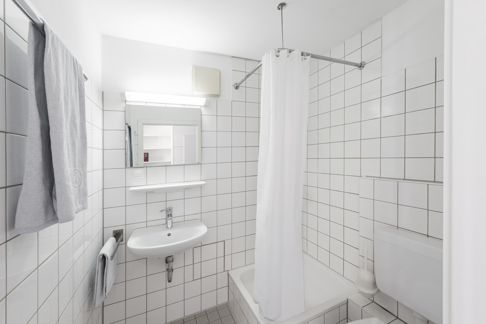 Badezimmer mit weiß gefliesten Wänden, Waschbecken, Dusche mit Vorhang und Toilette, Handtuch an der Stange.