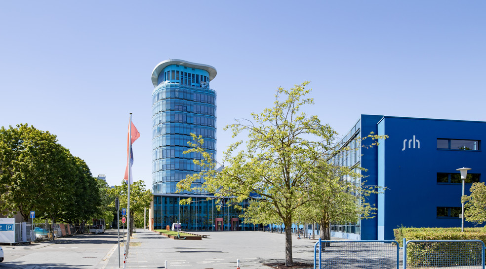Moderne Glashochhausanlage mit blauem Gebäude und Flagge auf Vorplatz, umgeben von Bäumen und Parkfläche.