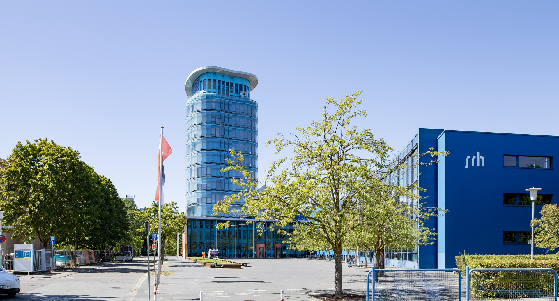 Moderne Glashochhausanlage mit blauem Gebäude und Flagge auf Vorplatz, umgeben von Bäumen und Parkfläche.