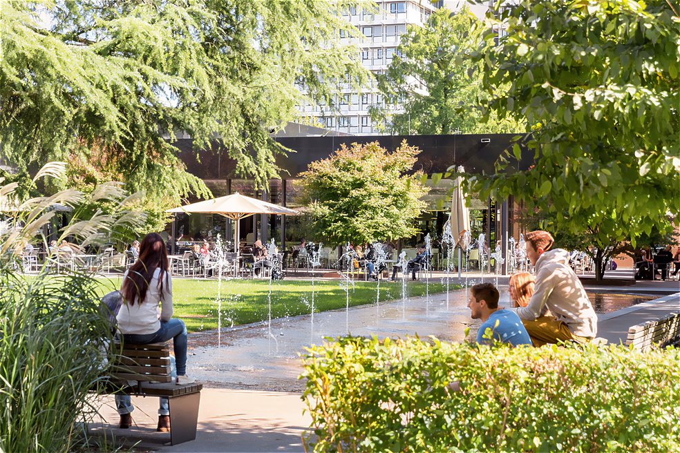 Menschen sitzen auf Bänken neben einem Brunnen mit Wasserspielen, im Hintergrund Café mit Gästen und Bäumen.