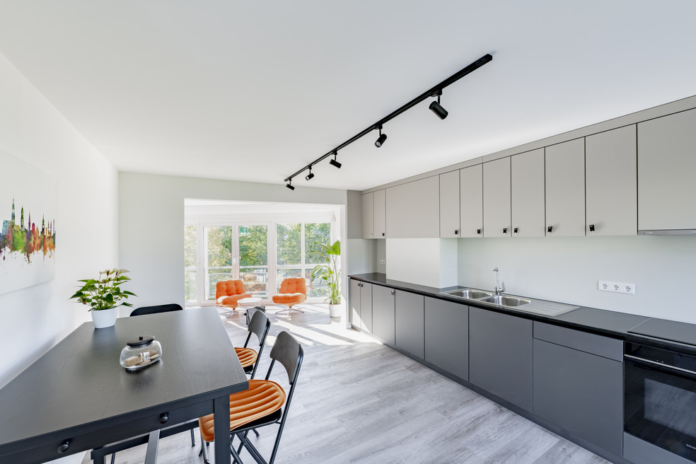 Moderne Küche mit Essbereich, hellen Schränken, schwarzem Tisch und orangefarbenen Stühlen, große Fenster mit Blick nach draußen.