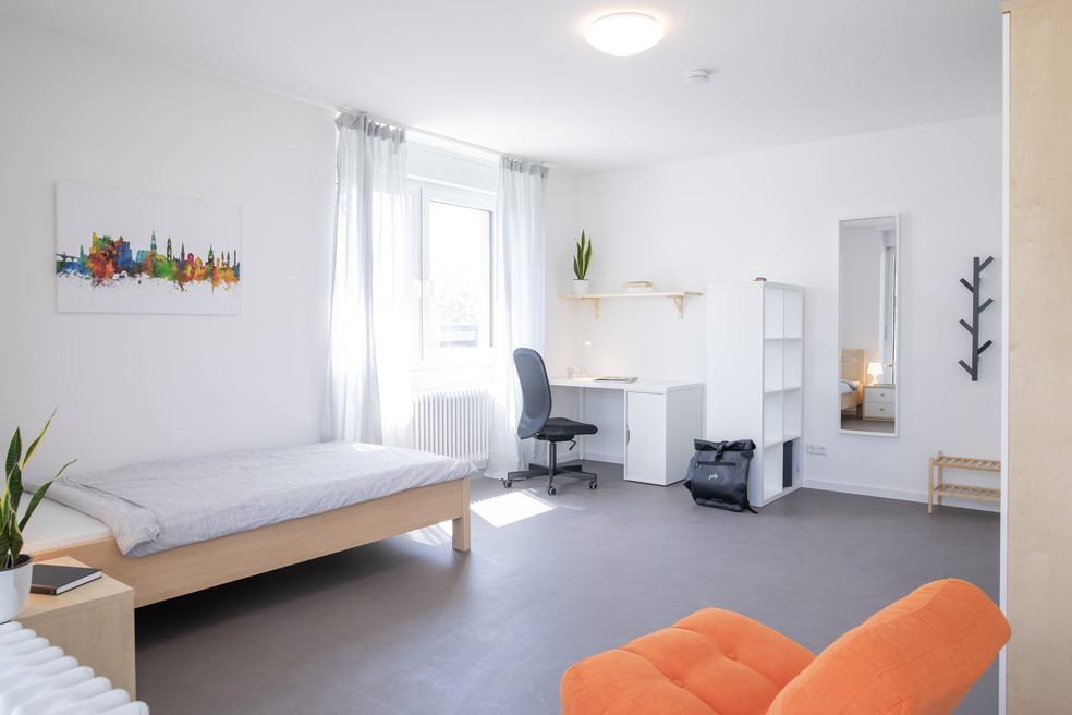 Ein helles Zimmer mit Bett, Schreibtisch, Stuhl, orange Sessel, Regal und Fenster mit Vorhang. Deko zeigt eine Stadt-Silhouette, daneben Spiegel und Pflanze.