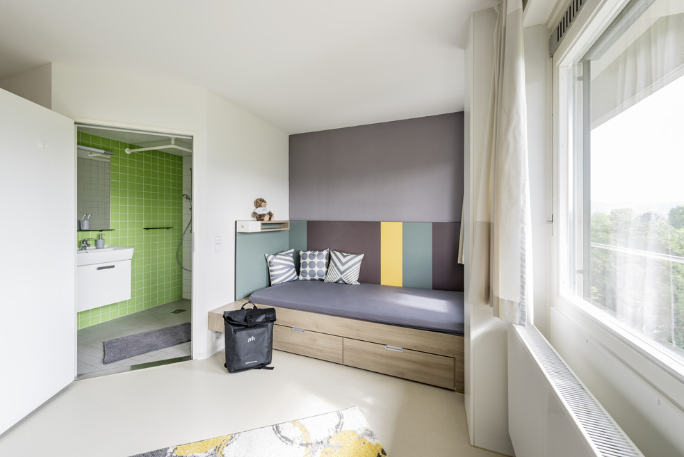 Kleines Zimmer mit Bettsofa, Schublade, Fenster und angrenzendem Duschbad in Grün. Koffer steht am Fußende, Teppich am Boden.