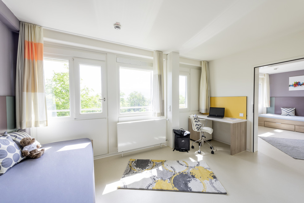 Moderne Wohnung mit Bett, Schreibtisch, Computer, Rollcontainer und großen Fenstern, helles Ambiente. Teppich und Sitzgelegenheit im Raum.