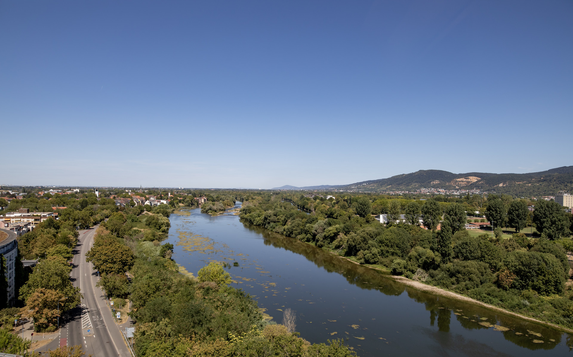 Luftaufnahme eines Flusses mit angrenzenden Wäldern, Straßen und Stadtteilen im Hintergrund.